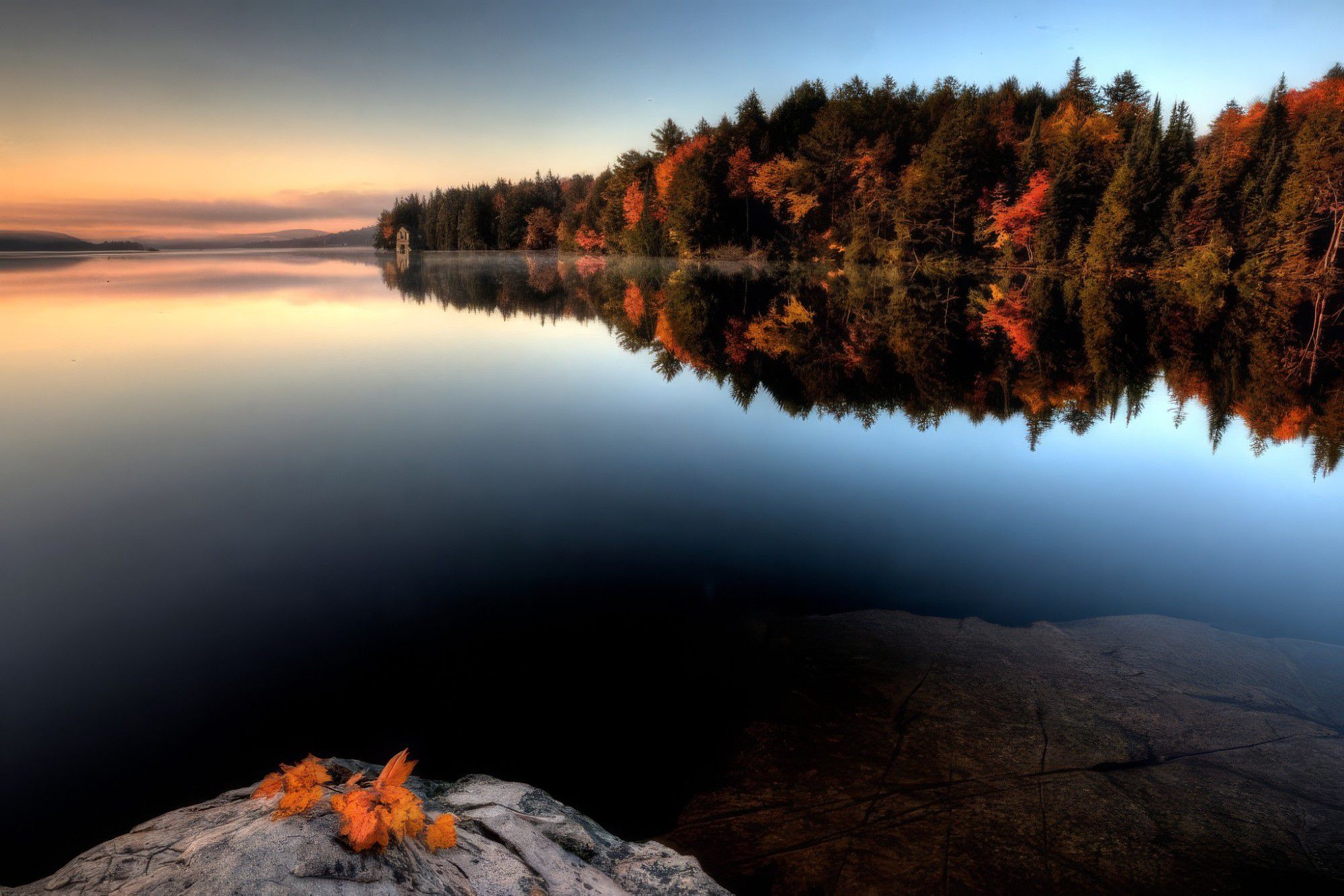 Still Ontario lake at dawn with autumn forest reflected in the water and maple leaves on a granite rock