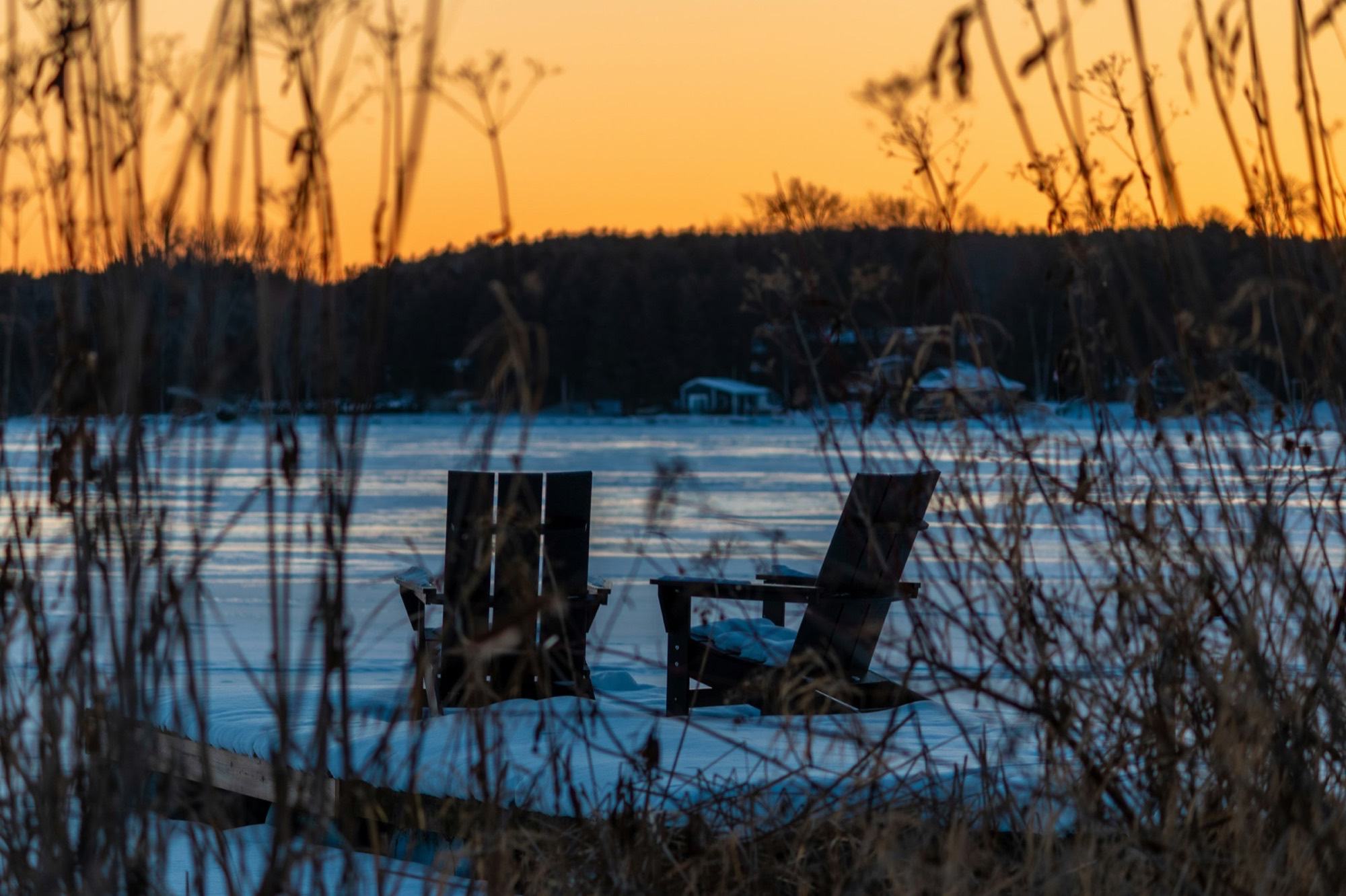 Two Adirondack chairs in the snow facing a frozen lake at sunset